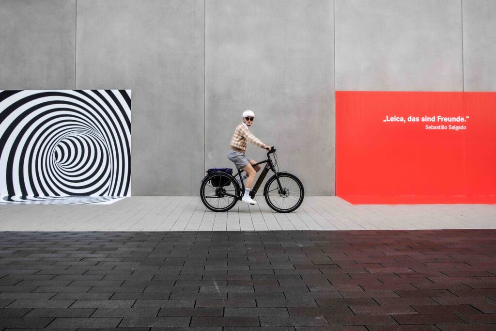 Frau fährt auf einem E-Bike, im Hintergrund ein Gemälde mit der Aufschrift "Leica, das sind Freunde"