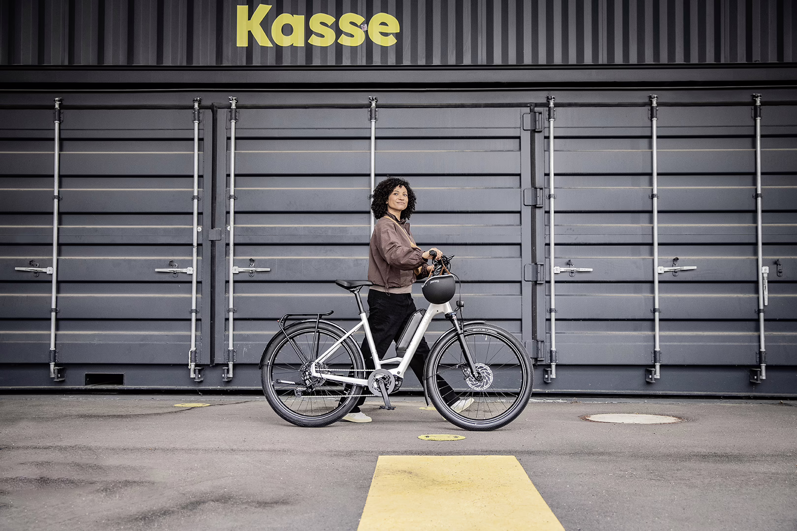 Eine Frau mit brauner Lederjacke schiebt ihr E-Bike vor einer Fassade aus metallischen Elementen, die an einen Container erinnern.