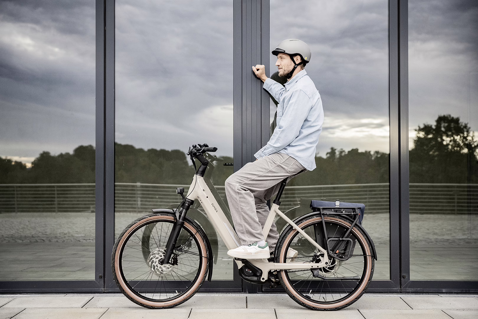 Ein Mann gekleidet im Casual-Stil sitzt auf einem E-Bike und stützt sich mit der Hand an einer Glasfassade ab