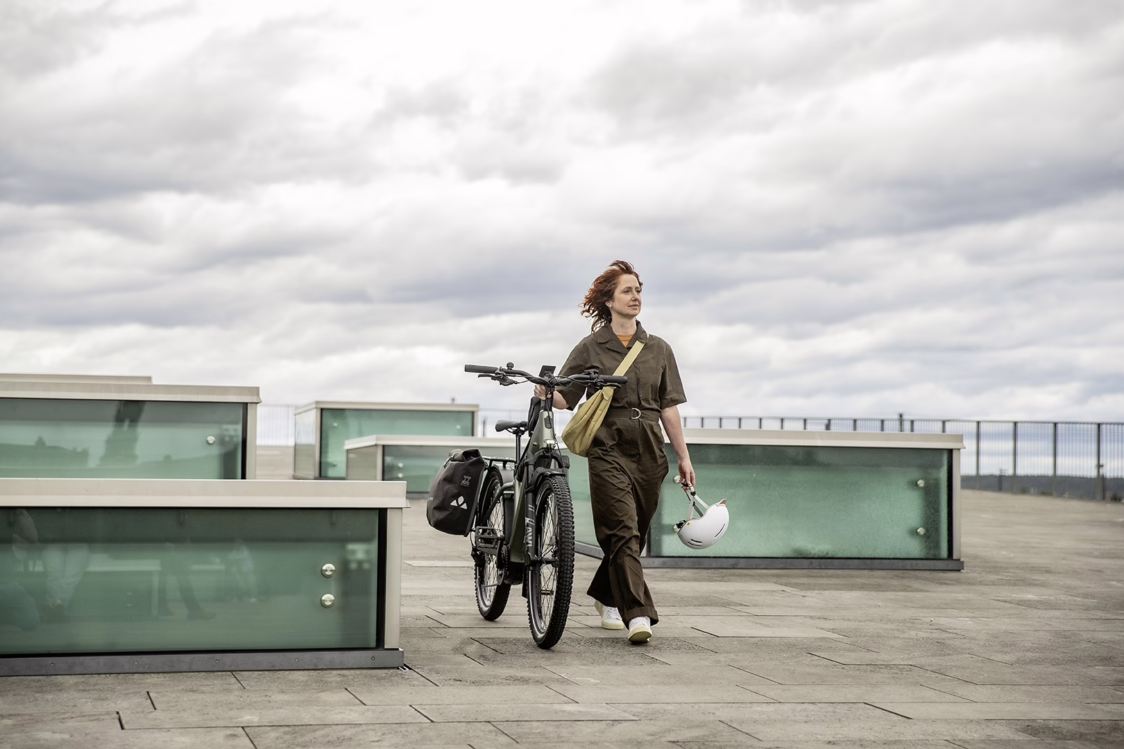 Eine Frau im grünen Overall schiebt ihr E-Bike über einen Platz mit modernen Architekturelementen