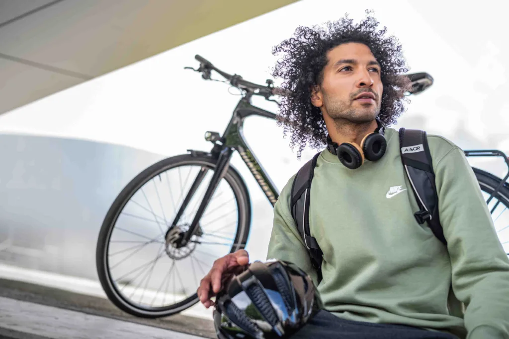 Mann mit Kopfhörern sitzt auf einer Mauer, im Hintergrund steht ein Fahrrad