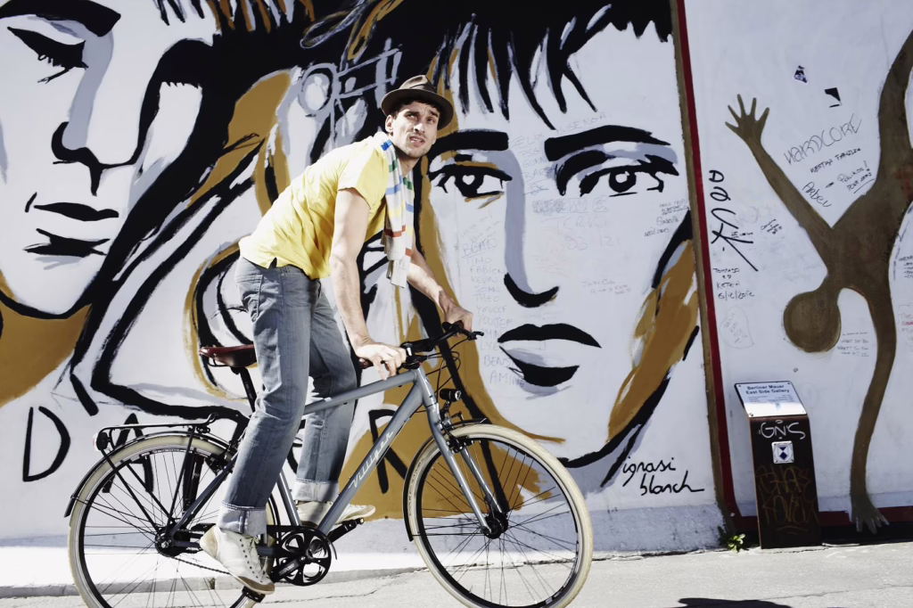 Ein Mann fährt vor einem Graffiti eines Mauer-Künstlers Fahrrad, er balanciert das Rad im Stehen, trägt ein gelbes T-Shirt und einen braunen Hut