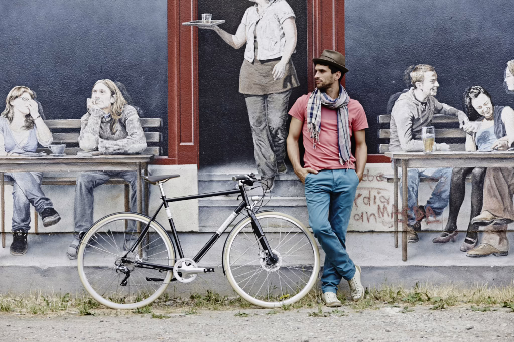 ein Mann in Jeans und lachsrotem T-Shirt mit Hut und grauem Schal steht vor einem Wandgemälde, das erwachsene Menschen an Tischen vor einem Haus zeigt. Neben ihm steht sein Diamant 130