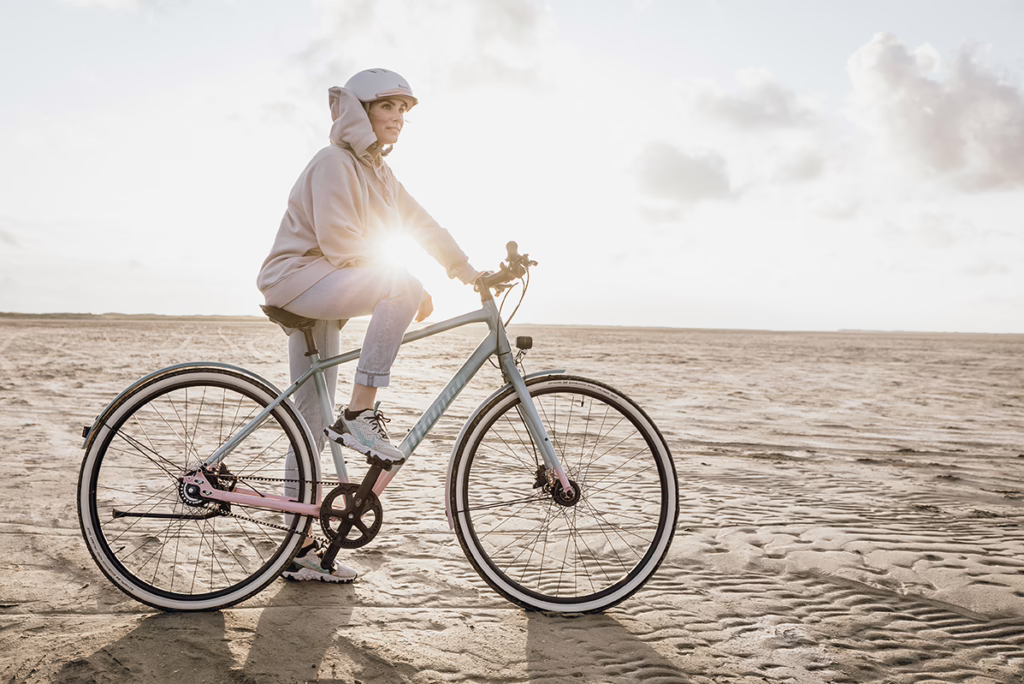 Eine Frau auf einem Strand an der Nordsee sitzt auf ihrem Fahrrad, das linke Bein am Boden, während im Hintergrund die Sonne aufgeht